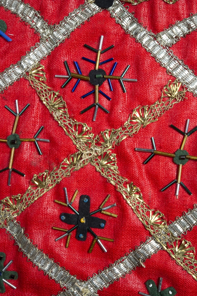 Rød kristnableie i ull med dekor av metallbånd, glassperler og klede.