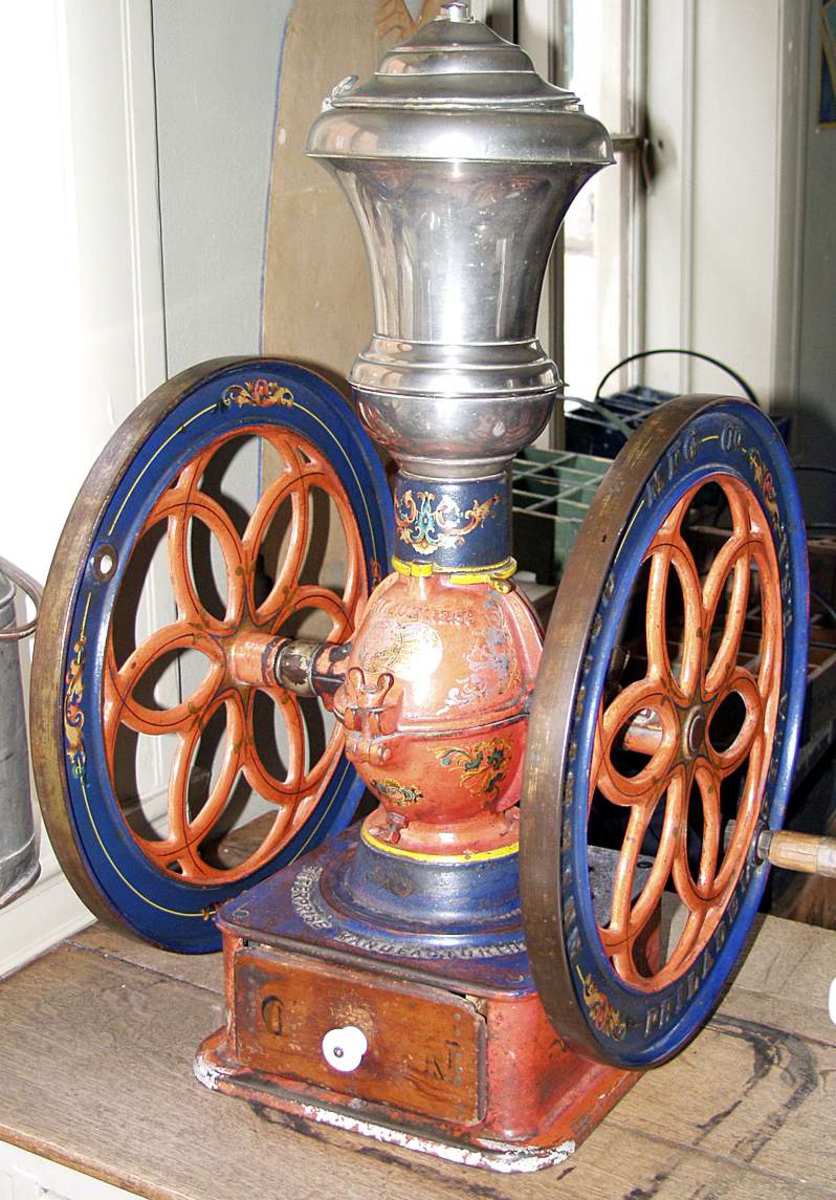 Kaffekvern i orangerosa støpejern med sveiv og to svinghjul. Trakt og beholder i blikk. Skuff og sveivehåndtak i tre.