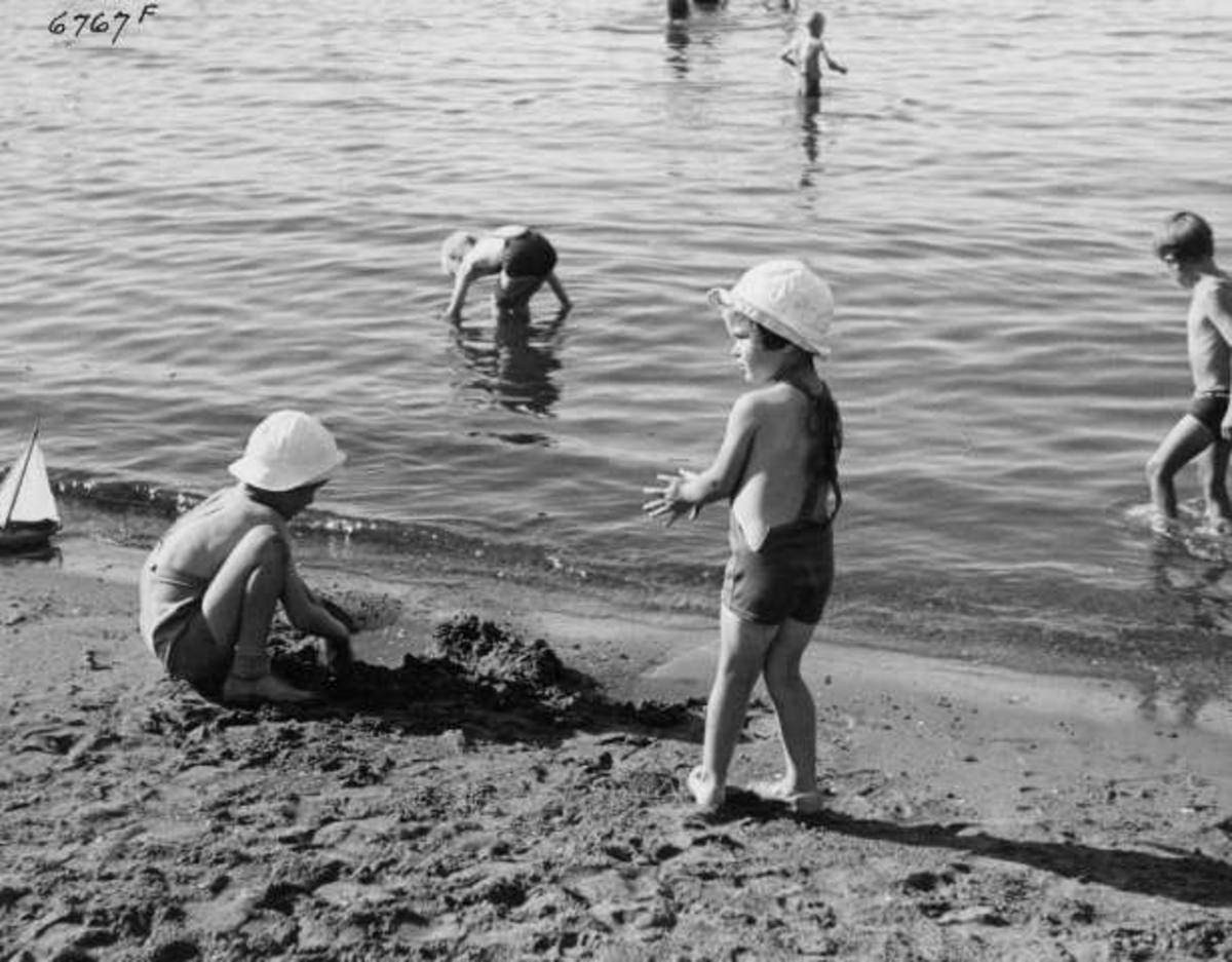Strandliv med badegjester  ved sjøen på Ingierstrand bad utenfor Oslo. Barn leker i sanden.