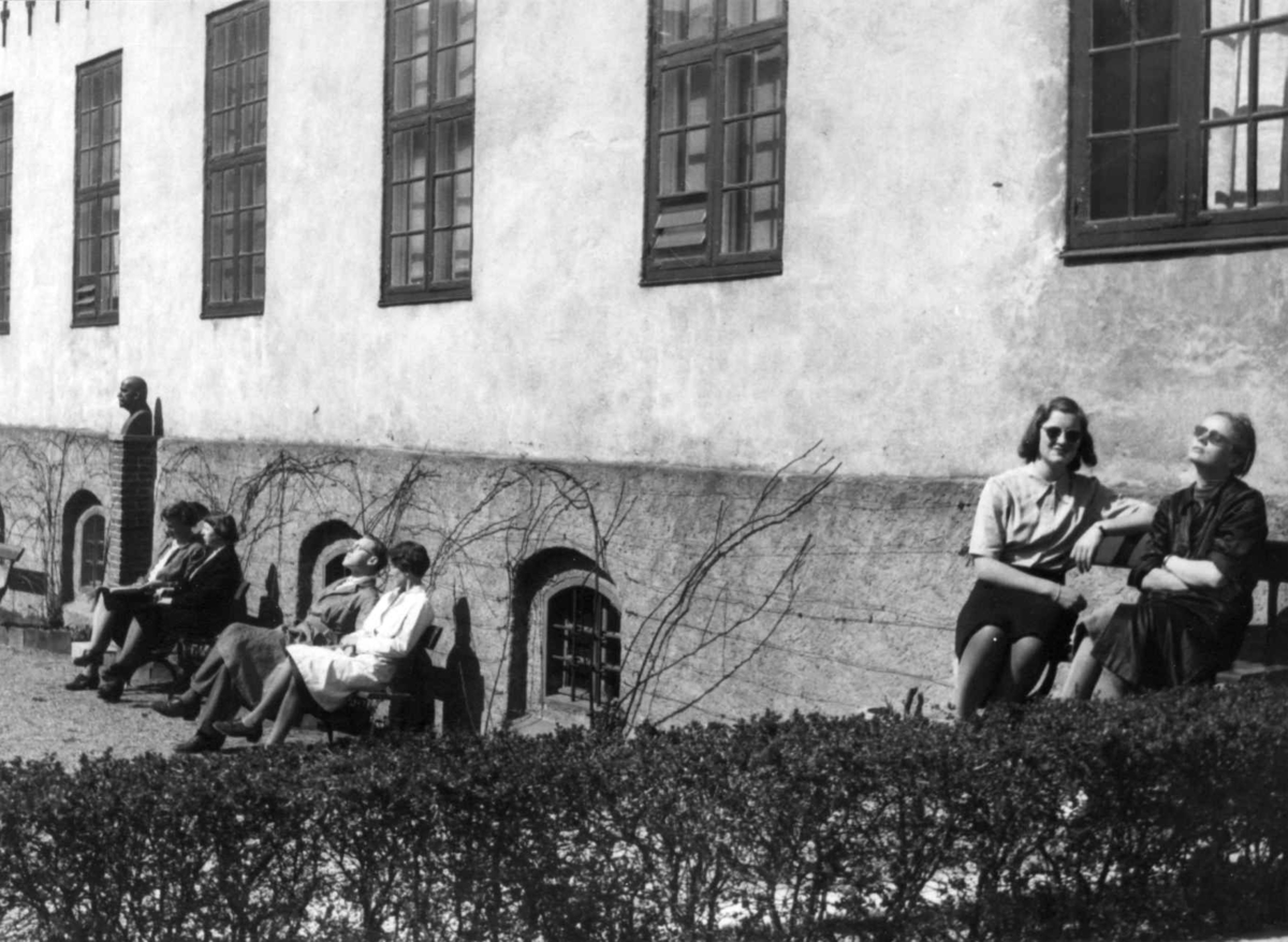 Flere av museets ansatte  på  benker i solveggen foran Bybygningen, bygning nr. 311, 1942.Bysten av Hans Aall i bakgrunnen. 