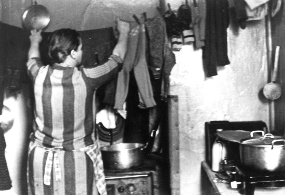 Interiør, kjøkken, Oslo. Kvinne henger opp tøy på snor over komfyr med gryter.
Fra boliginspektør Nanna Brochs boligundersøkelser i Oslo 1920-årene.