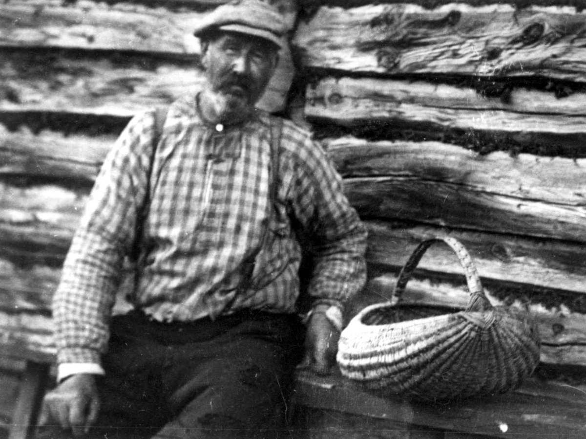 Ole O. Ålbu, bureisingsmann, sitter med en røverballkurv av kløvd bjørketau foran en tømmervegg. Oppdal, Sør-Trøndelag 1934.
