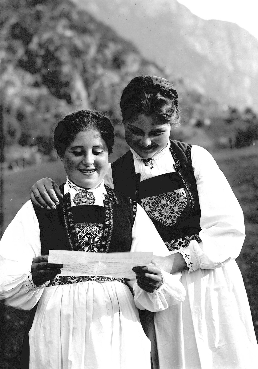 To kvinner i nasjonaldrakt holder rundt hverandre på jorde/eng. Den ene holder et brev som de leser.
