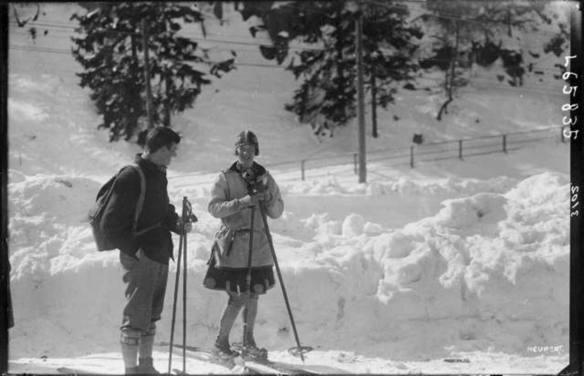 To skiløpere i samtale, mann med sekk og kvinne
i skjørt.