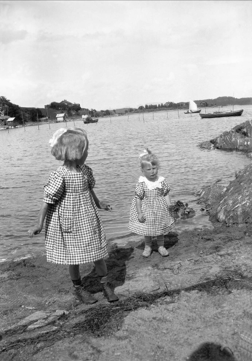 To jenter kledd i kjole står nede ved en strand. Robsahm og Lund.