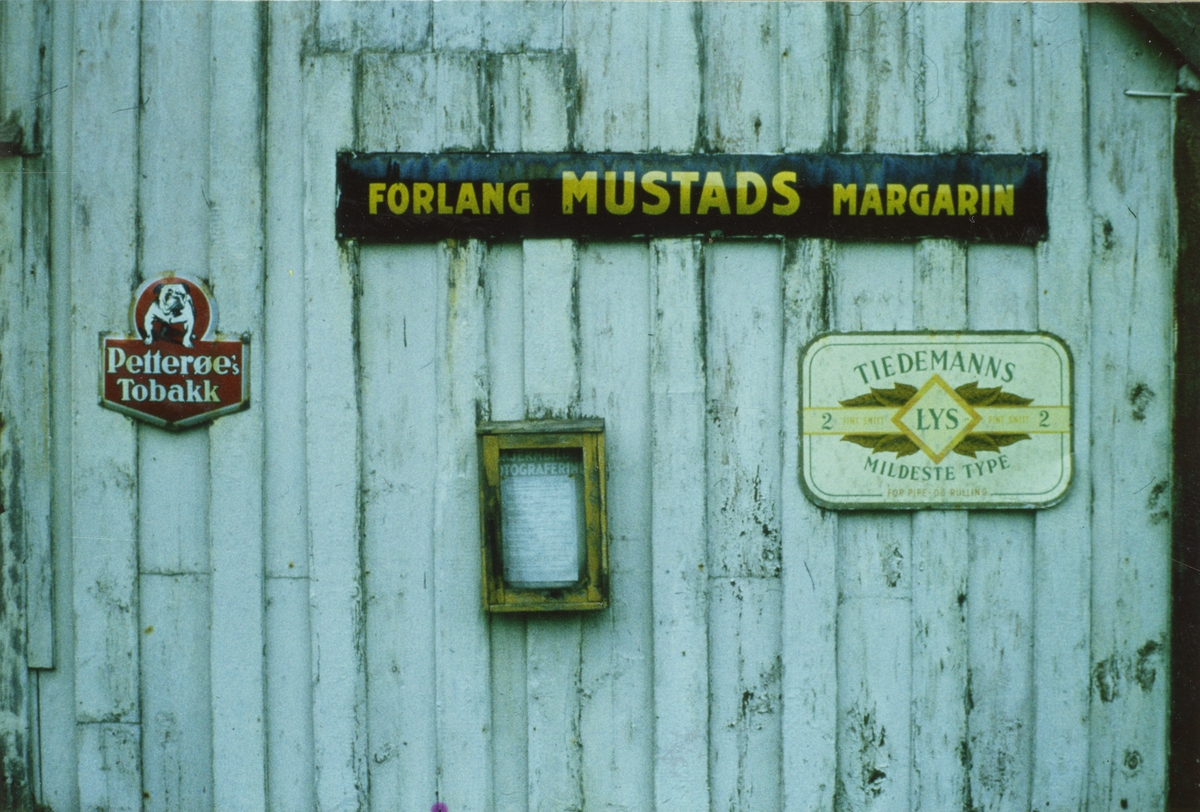 Husvegg med reklameskilt. Reklame for Mustads margarin, Petterøes Tobak og Tiedemanns Lys.