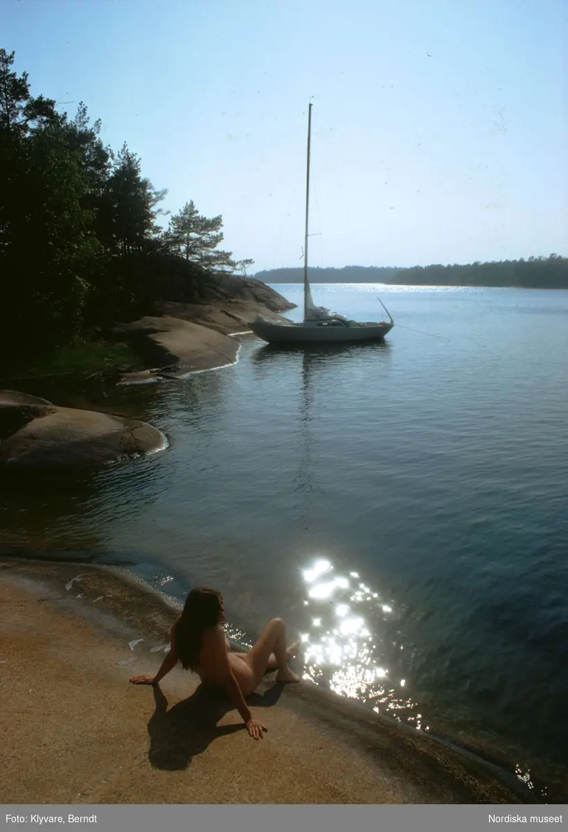 Naken kvinna på klippa i Stockholms skärgård. I fonden en segelbåt.