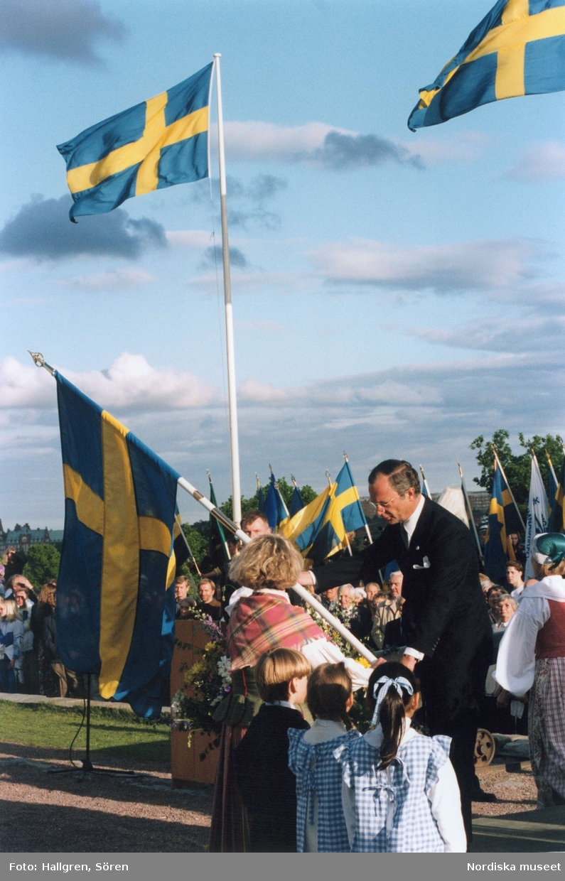Sveriges Nationaldagsfirande på Skansen, Svenska Flaggans dag. Kung Carl XVI Gustav delar ut en fana till kvinna folkdräkt.