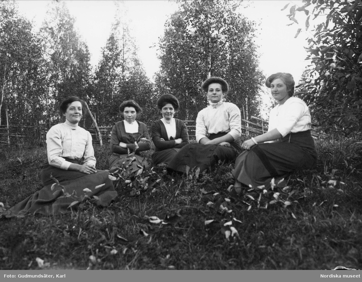 Fem kvinnor sitter i gräset. I bakgrunden gärdsgård.