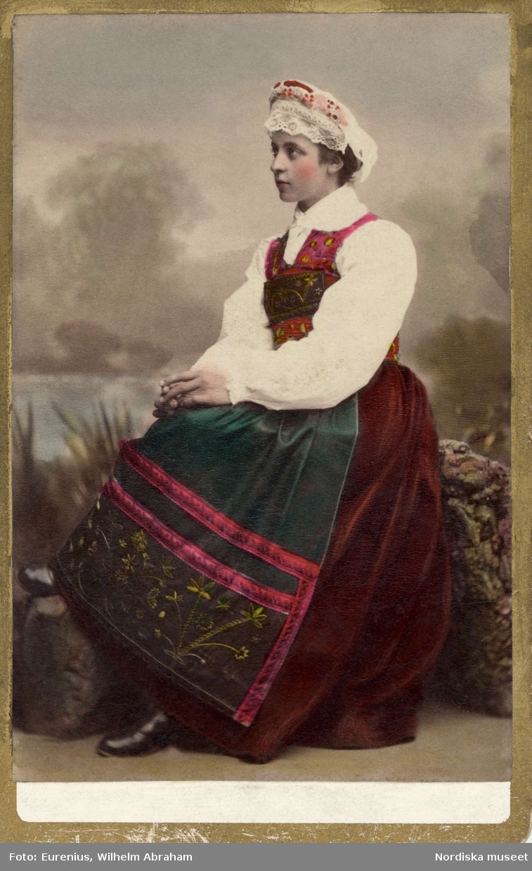 En kvinna i folkdräkt sitter vänd i profil. Högtidsdräkt från Österåker, Södermanland. Som den bars omkr. sekelskiftet 1900