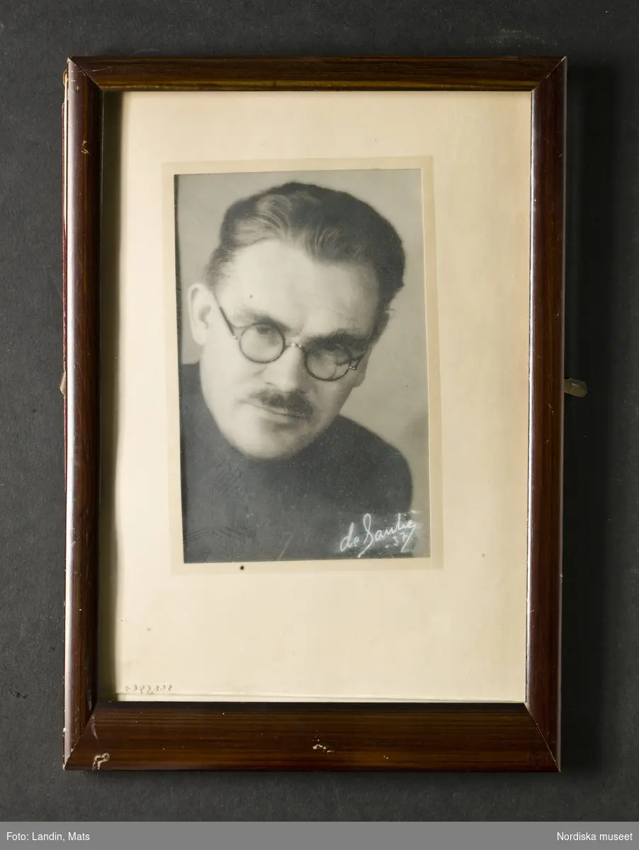 Porträtt, ansiktsbild, av man. Föreställer John Gran, 1937. Mustasch och små runda glasögon. Iklädd svart tröja. Fotografiet signerat i nedre högra hörnet: 'de Santié/-37'. Fotografi i ram. Nordiska museet inv.nr 258696.