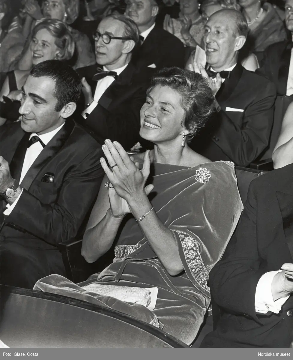 Porträtt av skådespelerskan Ingrid Bergman (1915-1982). Ingrid Bergman sitter i en publik med festklädda, applåderande människor. Själv är hon klädd i draperad, sten- och pärlbesatt sammetsklänning. Hon bär smycken med pärlor och stenar.