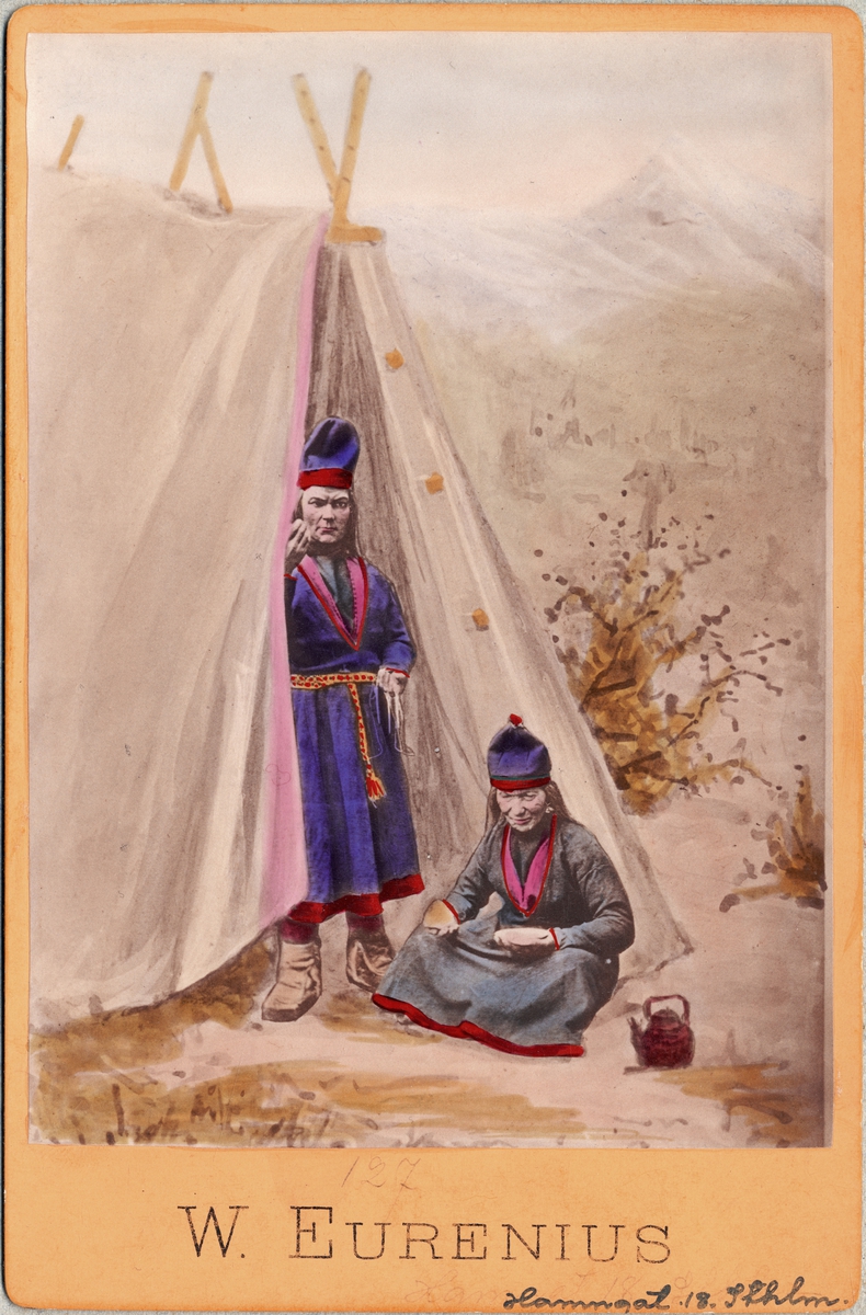 Dräktdockor föreställande två kvinnor i samiska dräkter vid kåta. Handkolorerat foto.