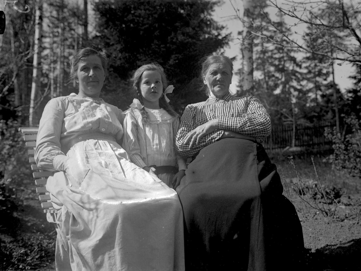 Gruppbild av två kvinnor och en flicka på en bänk. I bakgrunden ett staket.