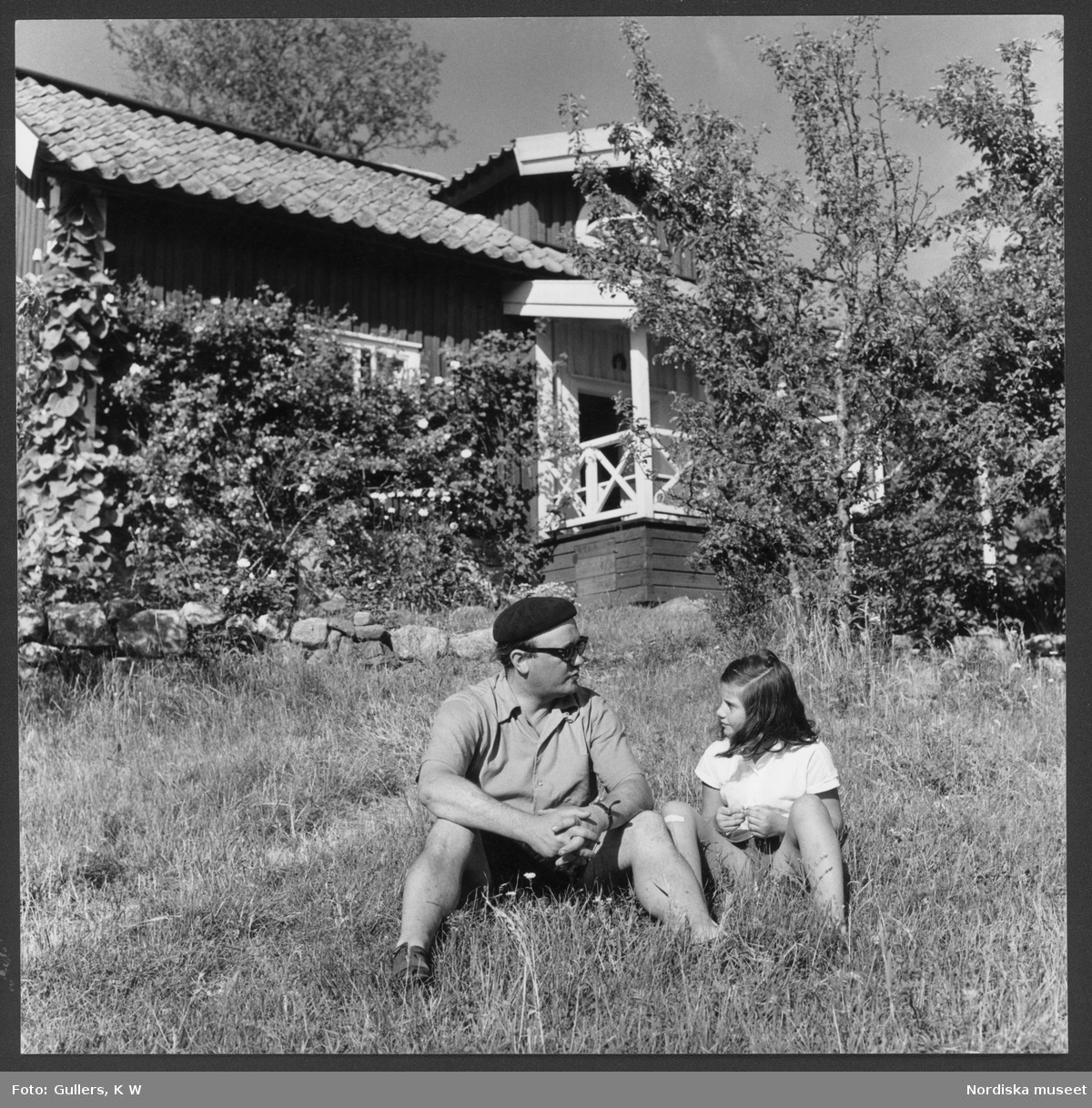 Stieg Trenter med dotter sittande i sommargräset. Hus i bakgrunden. Gåtebo, Gryts skärgård, Östergötland.