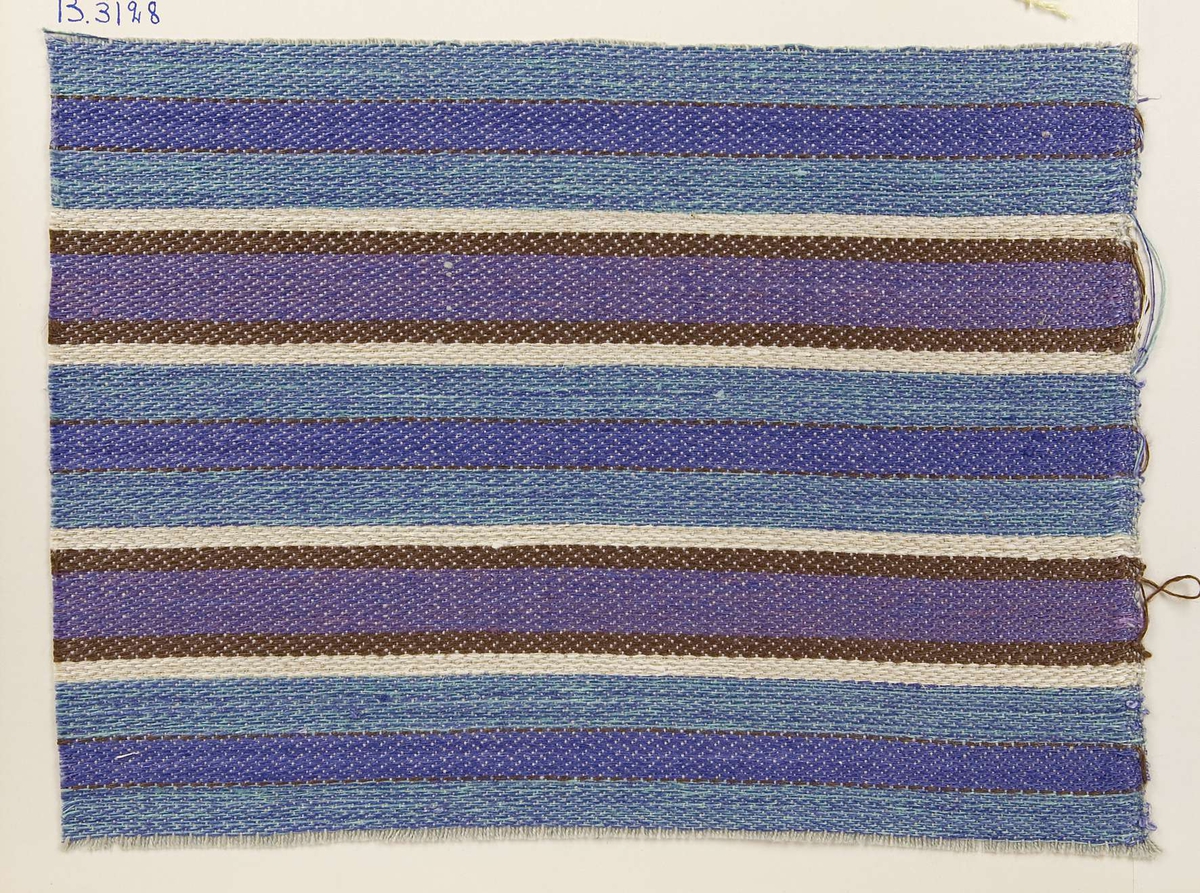 Vävprov ämnat för möbeltyg vävt med lin- och bomullsgarn, randigt i blått, lila, vitt och svart. Vävprovet har nummer "B-3128".