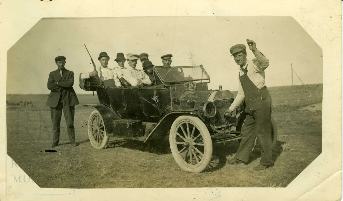 Seks personer sitter i en åpen bil. En mann står foran bilen og holder sveiva i ene handa. En mann står ved siden av bilen.