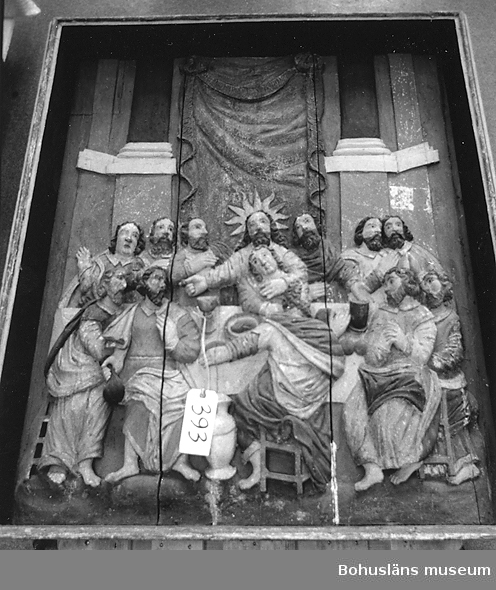 Altaruppsats från 1675, rikt skuren i trä med central skulpturscen, krön, sidostycken, pelare och kapitäl som krederats och målats med mager oljefärg samt förgyllts med oljeförgyllning. Förgyllningen uppvisar både bladguld, slagmetall och förmodligen silver, som mörknat påtagligt. Tillhörande skulpturer överst på ömse sidor om kröndekoren; UM394, UM395.

Centralmotivet skildrar nattvardens instiftande i en skulpturscen. Över scenen läses nattvardens instiftelseord på danska.
Under skultpruscenen inskriften: 
ANNO 1671 ER DENNE KIERKEBYGNING I JESU NAFN BEGYNT OCH UNDER PASTORIS JOANNIS COLSTRIPII FLID MEDH SIT INREDE ANNO 1675 FULBORDET MEDH MIDDEL FRA EGNE RINGE INTRADER, ANDRE GOT FOLCKIS TILHJELP OCH FORNEMMELIG H. KONGL. MAJIT:S WOR ALLERNADIGSTE KONUNG CAROLI XI TILGAWE AFF 150 RIXDALER.

Altartavlan komer från Norra Ryrs kyrka, riven 1873 och benämnd Norra Ryrs kyrka fram till år 1884, idag Lane-Ryrs kyrka. 
UM000384, UM000385, UM000389a-i, UM000393a-i, UM000394, UM000395a-b, UM000401, UM000404a-b, UM000405a-g, UM005660:1-57 samt UM65.01.001 är samtliga inventarier från den gamla kyrkan.

Beskrivning av altaruppsatsens delar:
a. Tavla med nattvardsframställning; h. 120, br. 104 cm
b. Överdel; l. 224 cm
c. Krön till överdel; br. 132, h. 58 cm
d. Pelare; h. med platta över kapitälet 113 cm
e. Pelare, som föregående h. 113 cm
f. Sidostycke; i 2 delar I-II, h. med tappar 128, br. 67 cm
g. Sidostycke; i 2 delar I-II, h. med tappar 131, br. 69,5 cm
h. Underdel; l. 201 cm
i. En lös del, tillhörande UM000393h

Altaruppsatsen ska enligt uppgift i artikel i Bohusläningen den 30 april 1963 vara skänkt till församlingen av Karl XI som ett led i försvenskningen av Bohuslän.

Ytterligare foto i Uddevalla museums kyrkliga utställning år 1920, se UMFA53292:0013, UMFA53710:0075.

Ur Nationalencyklopedin, NE.se: Altarprydnad
Under 1500-talet utbildades i Italien en ny typ: en central målning eller skulptur, omgiven av en rik, klassiserande omramning. I den tyska renässansen utvecklades altarprydnaderna till altaruppsatser, som med kvardröjande gotisk höjdsträvan byggdes upp i flera våningar. Centrala bilder, målade eller skulpterade, av (nedifrån räknat) nattvarden, korsfästelsen, uppståndelsen och himmelsfärden omges av en arkitektonisk ram med slösande rik ornamentik, allegoriska figurer av dygderna, änglar med pinoredskap etc. Denna typ blev vanlig i Norden under 1600-talet och utformades t.ex. av Jacob Kremberg, Lund, och Marcus Jäger, Göteborg.

Ur handskrivna katalogen 1957-1958:
Altartavla, Lane Ryr
a) Tavlan m. nattvardsframställn. H. 120; Br. 104 cm. b) Överdel, L. 224 cm. c) Krön t. överdel, Br, 132; H. 58 cm. d) Pelare, H. (m. plattan över kapitälet) 113 cm. e) Pelare, som  föreg. H. 113 cm. f) Sidostycke, H. (m. tappar) 128 cm; Br. 67 cm; i 2 delar I-II. 
g)  Sidostycke, H. (m. tappar) 131; Br. 69,5 cm; i 2 delar I-II. h) Underdel, L. 201 cm. i) En lös del, tillhörande U.m 393 h.

Lappkatalog: 13

Litt.: Gardell, Sölve: Den kyrkliga konsten i Uddevalla museum. Vikarvet 1933. 
Altaruppsats från 1600-talet åter i bruk i Lane-Ryrs kyrka. Artikel i Bohusläningen den 30 april 1963.
Kristiansson, S.: Uddevalla stads historia, del I, s. 338-339.
Oedman, Johan Chorographia Bahuensis. Thet är Bohus-Läns Beskrifning. 1746. Faksimil- tryck 1911, s. 110.