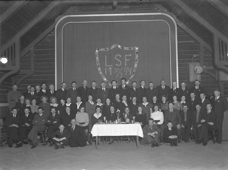 Text till bilden:"Män och kvinnor i en stor sal. Deras föreningsmärke på väggen. Framför dem ett bord med polaler. L.S.F 19 30/3 44".