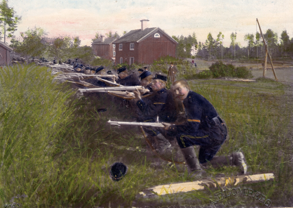 Soldater ur Andra Svea artilleriregemente A 5 ligger skjutklara med gevär i skyttelinje vid bondgård(?). Civila åskådare ser på i bakgrunden. Färglagt foto från 1902.