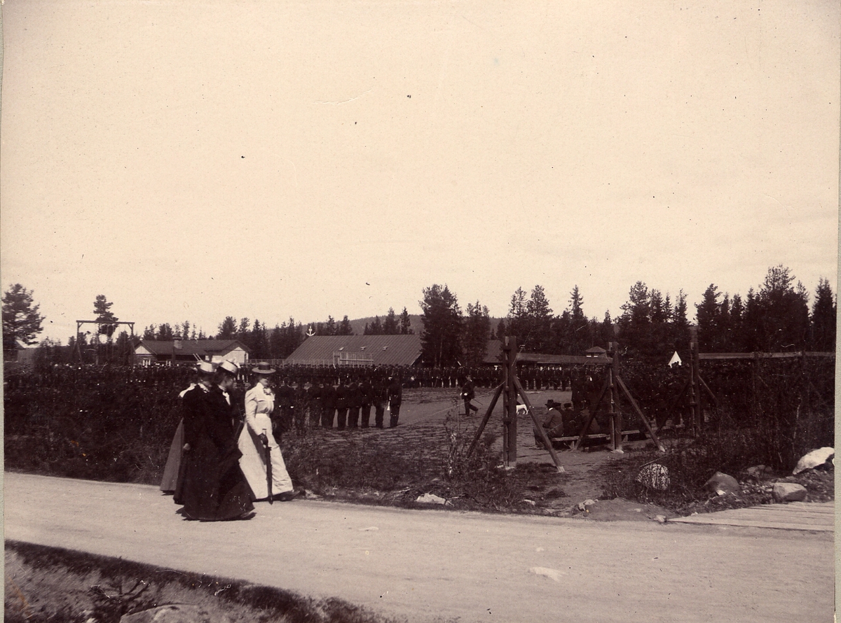 Soldater uppställda för gymnastik på kyrkoplan i Sollefteå läger. I förgrunden fyra kvinnor på promenad.