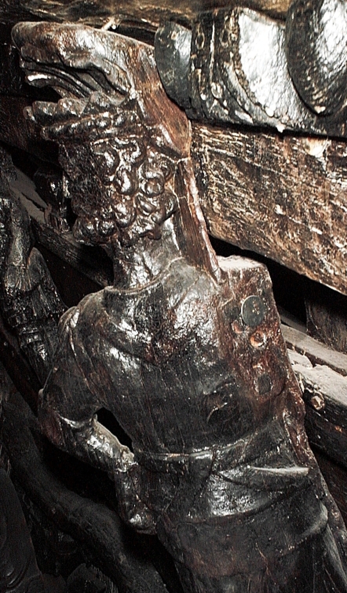 Skulptur föreställande en mansfigur iförd romersk rustning. På huvudet har figuren en taggig krona med bred "stenbesatt" kronring. Bakom huvudet syns ett stort snäckskal. Ansiktet pryds av ett stort, relativt kortklippt och krusigt skägg. Figuren är iförd en långärmad rustning med stålharnesk, axelskydd och knäskydd. Rustningen är, som typ betraktad, ett mellanting mellan romersk rustning och renässansrustning.



Figuren står vinkelrätt mot bakstycket, med höger sida tryckt mot bakstycket och vänder följaktligen den vänstra sidan utåt. Kejsaren har huvudet vridet kraftigt åt vänster så att han blickar mot betraktaren när denne står framför skulpturen. Den vänstra handen vilar mot höften. Höger arm, som saknas, har troligtvis hållits bakåt, bakom ryggen. Figuren vilar på höger ben, medan vänster ben hålls böjt. Möjligen avser framställningen att återge kejsaren i rörelse framåt, med tanke på att kroppen är framställd i profil och inte en face. Figuren står på en sockel, vilken pryds av en grinande maskaron samt en namninskription.



Upptill är bakstycket framdraget i en enkel "huvudplatta", med en delvis kvartscirkelformad profil. Skulpturens baksida uppvisar tre snedställda, olika djupt liggande fält. De tre fälten, som utgör anliggningsytor, ger skulpturen en trappstegsformad profil.

Sockeln är skadad nedtill och namninskriptionen går ej att tyda. Figurens högra arm saknas. I övrigt är skulpturen välbevarad.



Text in English: A sculpture representing a male figure wearing a Roman armour. On the head there is what looks like a flat crown and on the board behind the head there is possibly a large sea shell. The face has a large curly and mostly clipped beard and is framed by curly hair. The figure wears long-armed armour with a steel curass, shoulder guards and knee guards of a type reminiscent of both Roman and renaissance amour.



The figure stands at a right angle to the back-board of the sculpture, with its right side pressed to the back-board and the left side outermost. The figure''s head is turned to the left lookimg over his shoulder. The left hand rests on the hip were there originally would have been a sword. The right arm, which is missing, was held behind the back of the figure. The body rests on the right leg, the left leg is bent, the pose is possibly meant to show the figure moving forward. The figure stands on a plinth decorated with a mask and a name inscription.



The upper part of the back extends obliquely upwards in a simple"head board".



The back has three obliquely placed surfaces of different depths, that are areas of attachment. The surfaces make the sculpture looks like a step-ladder in profile.

The plinth is damaged and no letters on the inscription is visible. The figure''s right arm is missing. Otherwise, the sculpture is well preserved.