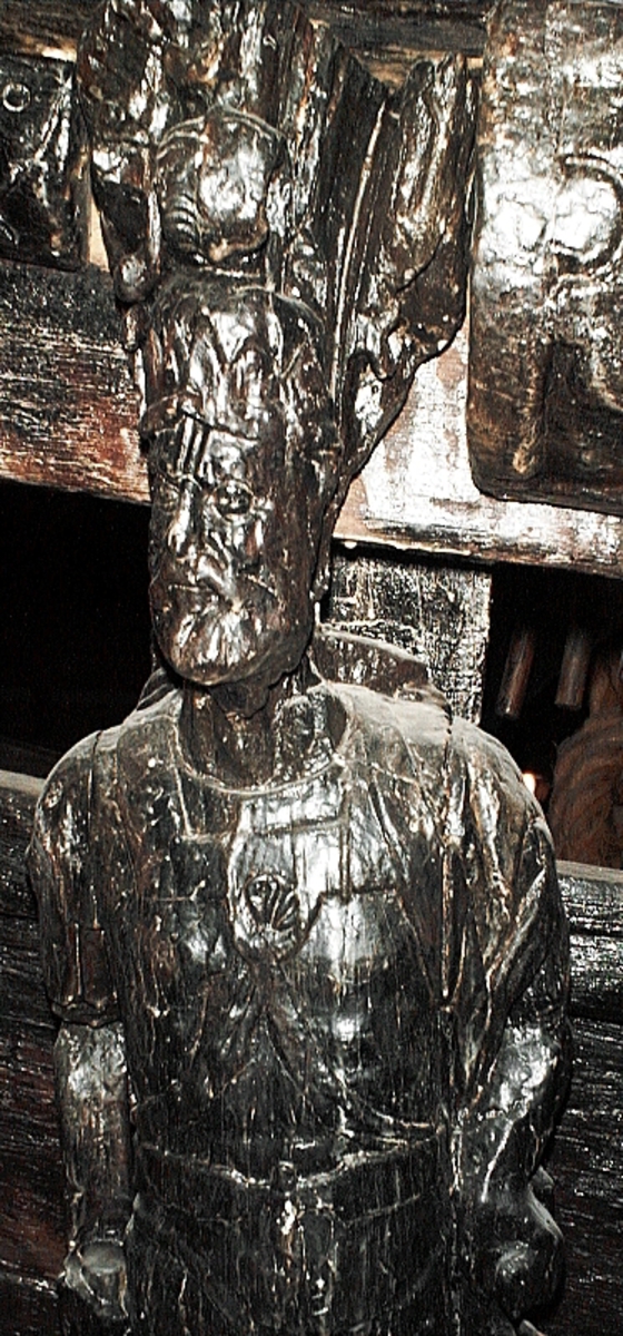Skulptur föreställande en mansfigur med ett långsmalt och skäggigt ansikte, under en hög, slät hjälm med stora plymer. Runt hjälmen är en stålkrona trädd. Bakom huvudet skymtar ett snäckskal. Figuren är iförd en rikt ornerad rustning av thoraxmodell. Utefter figurens rygg hänger en mantel, som också draperar den vänstra axeln.

Hela figuren är frontalt framställd. Höger arm hålls lätt böjd utefter sidan, med handen fattande om ett svärdsfäste. Vänster arm är något böjd rakt ut från sidan, och handen verkar fatta tag om manteln. Kroppstyngden är jämnt fördelad på båda benen.

Figuren står på en sockel, vilken pryds av en bred och grimaserande maskaron. Sockeln är även försedd med en banderoll som tycks helt slät och utan namninskription.

Upptill är bakstycket framdraget i en kraftig "huvudplatta". Skulpturens baksida uppvisar tre snedställda, olika djupt liggande fält. Fälten, som utgör anliggningsytor, ger skulpturen en trappstegsformad profil.
Huvudplattan är delvis skadad. Svärdets klinga saknas. I övrigt är skulpturen relativt välbevarad.

Text in English: A sculpture representing a male figure with a long and narrow face that is bearded and has a moustache. On the head there is a plumed helmet on which a crown with points has been placed and behind the head a large sea shell. The figure is wearing richly decorated thorax type armour. The mantle is hanging down the back and draped over the left shoulder.

The figure faces towards the front. The right arm is slightly bent and held at the side and the hand holds a sword hilt. The left arm is slightly bent and held out from the side and the left hand seems to hold the mantle. The weight of the body is carried on both legs.

The figure stands on a plinth with a broad and grimacing mask and also has a label which is quite smooth and without any inscription.

The upper part of the back extends obliquely upwards in a powerful "head board". The sculpture''s back has three obliquely placed surfaces of different depths that are areas of attachment. The surfaces make the sculpture look like a step-ladder in profile.
The board behind the head is partly damaged and the blade of the sword is missing but otherwise, the sculpture is rather well preserved.