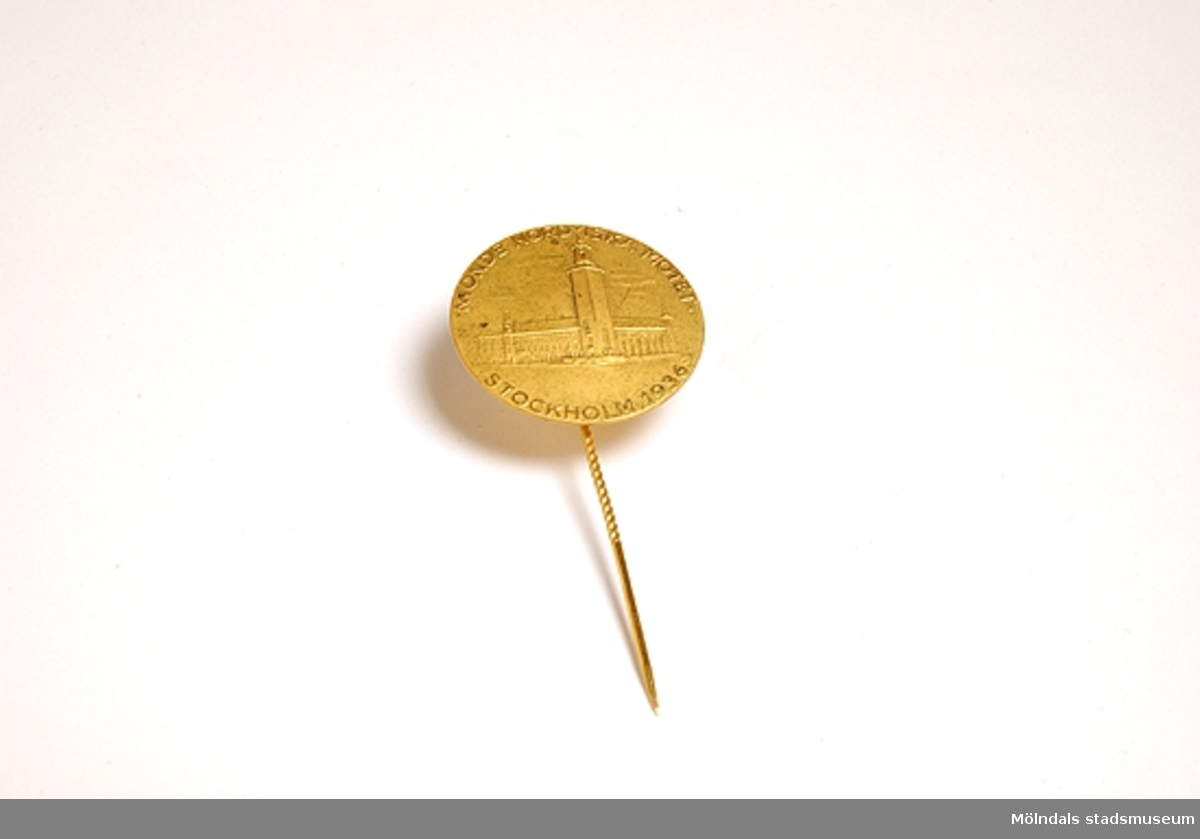 Ett runt märke i gulmetall med en nål, att fästa på ett jackslag. på märket är präglad en bild av Stockholms stadshus och texten Nionde nordiska mötet, Stockholm 1936.Givaren fann denna nål bland hennes mors efterlämnade saker.