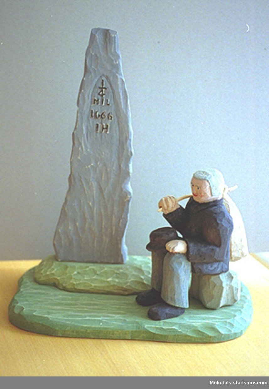 En äldre man sitter på en sten. Han håller en svart hatt i ena handen och en kappsäck över axeln i den andra. Vid sidan av står ett hög spetsig sten med inskriptionen: "1/4 mil 1666 IH". Harry Bergmans "gubbar" (träfigurer).
