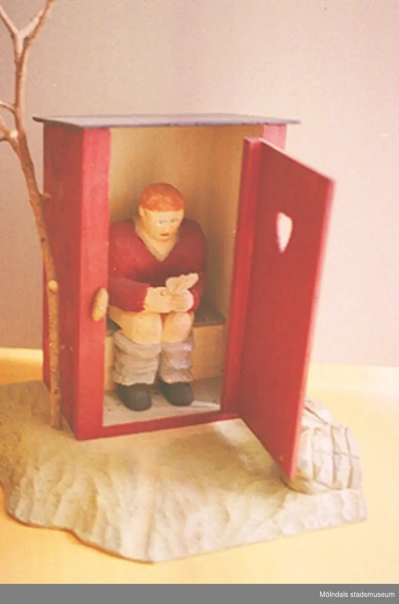 Ett rött utedass med öppen dörr. Därinne sitter en rödhårig person på toaletten och läser i en tidning. Den öppna dörren är försedd med ett hjärta. Utanför står ett litet träd. Harry Bergmans "gubbar" (träfigurer). Liknande foto som 1994_0964.