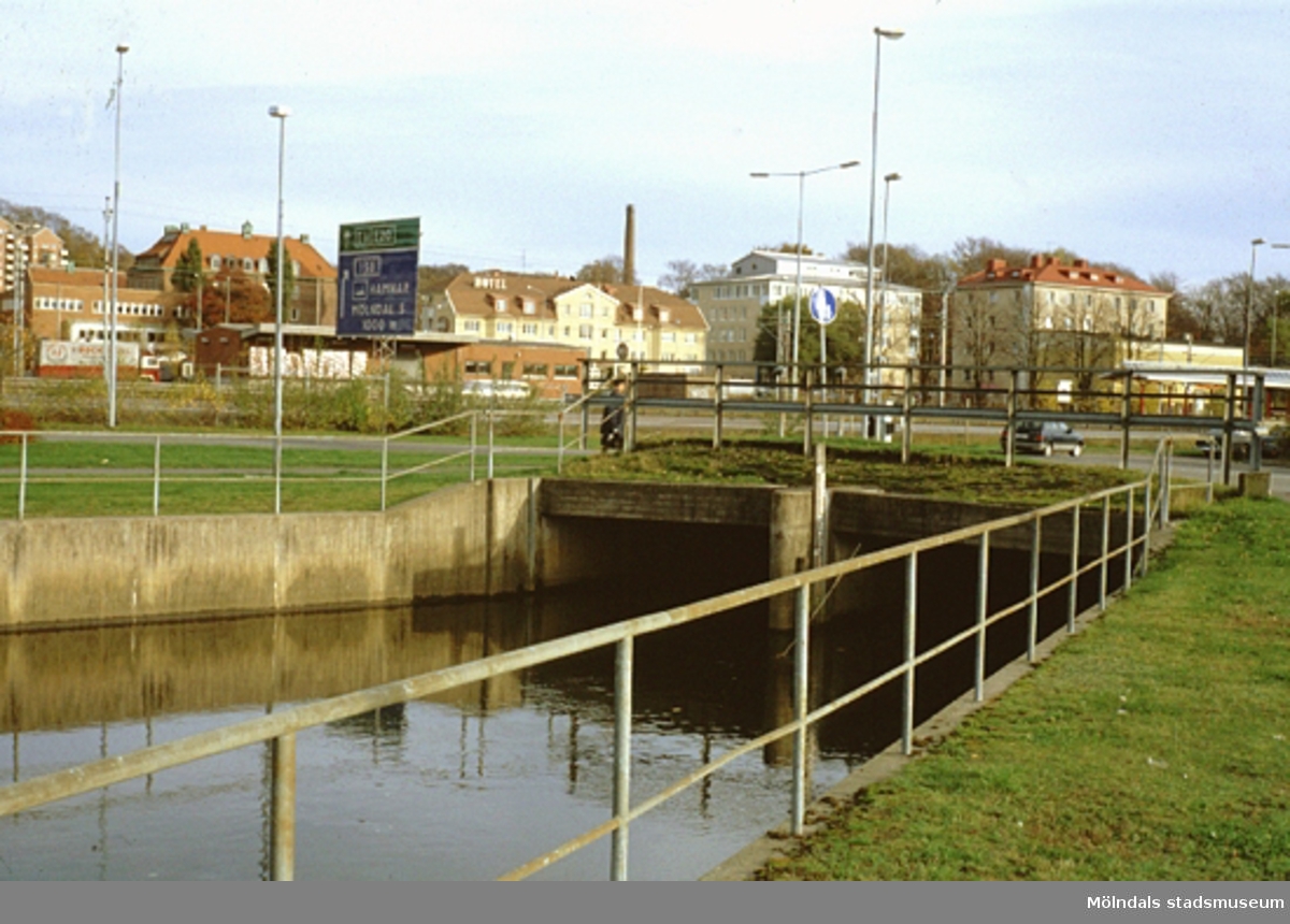 Kulvert i Mölndalsån utmed Göteborgsvägen, oktober 1993. Vy från Göteborgsvägen snett över E6/E20 mot sydost. I bakgrunden från vänster ses bl a Mölndals brandstation, Kvarnbyskolan, Hotel Mölndals Bro samt två kontorsbyggnader.
