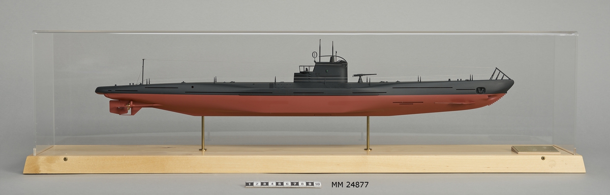 Ubåtsmodell Delfinen II i monter. Modell av alträ med detaljer av mässing, målad med cellulosafärg. Rött och svart skrov. Monter av plexiglas på träplatta. Mässingsbricka i montern med uppgifter om modellen.