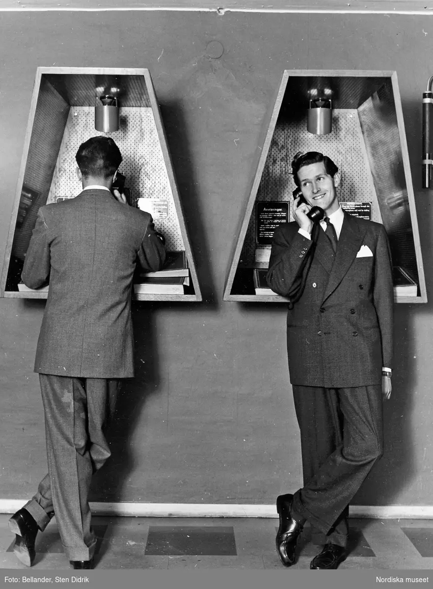 Två manliga modeller i kostym vid varsin telefonautomat.