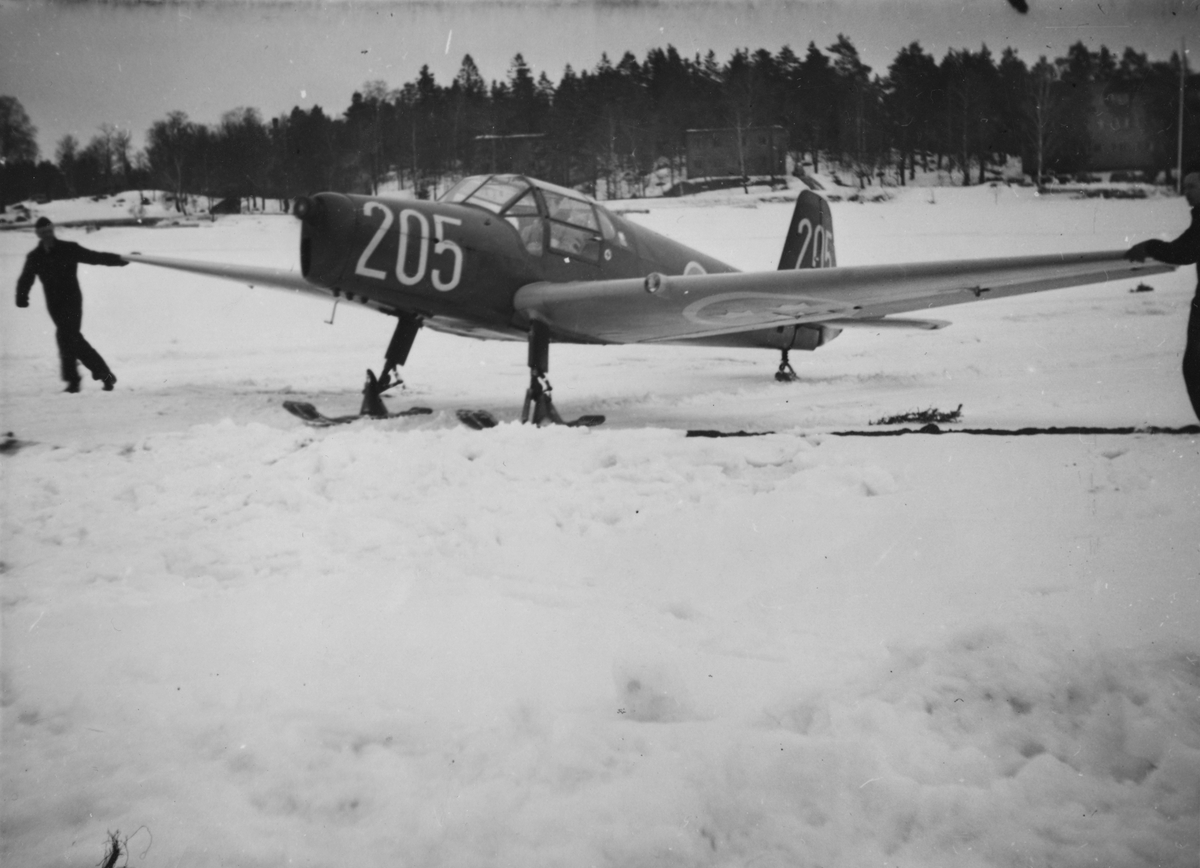 Flygplan SK 25 nummer 205 vid landning på flygfält på F 8 Barkarby, vintertid. Personer i arbete vid flygplanet.