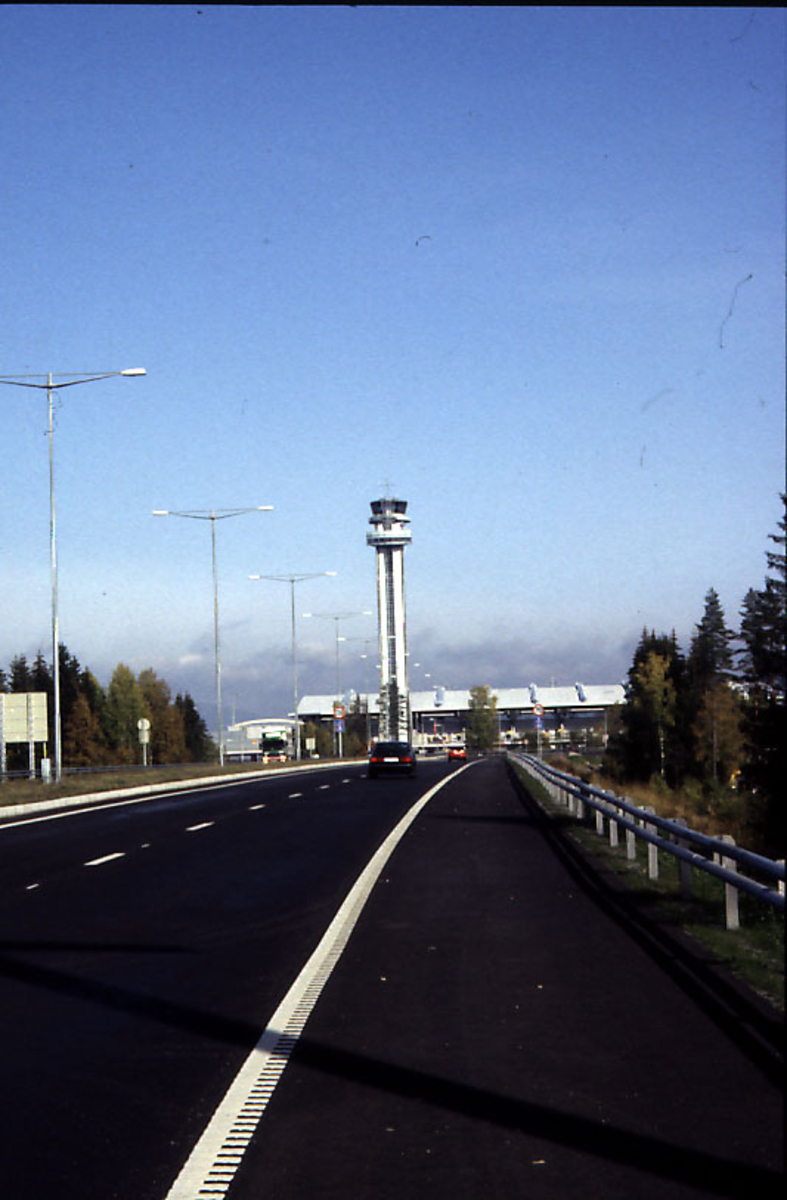 Lufthavn, vei som fører inn til lufthavnområdet.  3 kjøretøy på veien i forgrunnen.  Kontrolltårnet og terminalbygningen i bakrgrunnen.