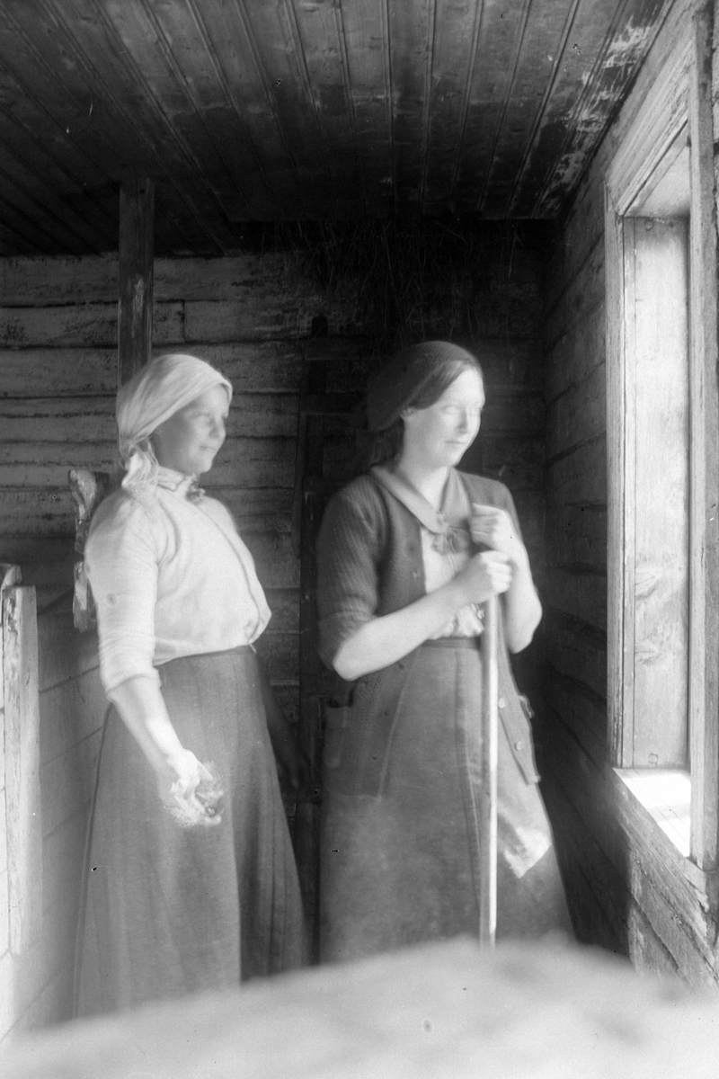 To kvinner fotografert innendørs i en laftet bygning, begge med skaut på hodet.