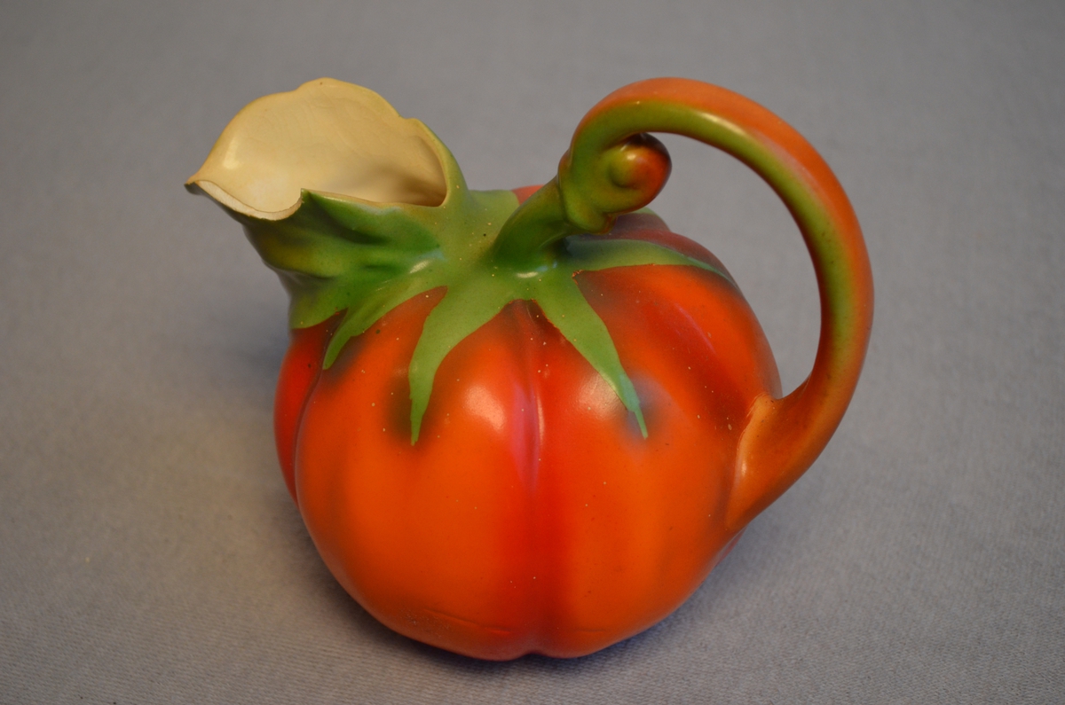 Mugge forma som ein tomat eller eit gresskar (?). Ein del av tuten er broten av. Den ligg nedi mugga.