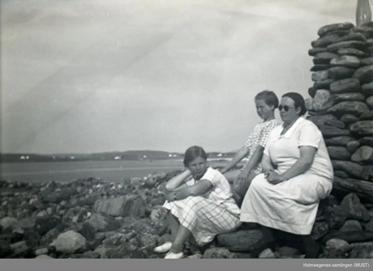En kvinne og to jenter kledd i sommerkjoler sitter ved en steinvarde. Antatt samme sted og tidspunkt som ST.K.HE 2007-011-0072 til -0081.
