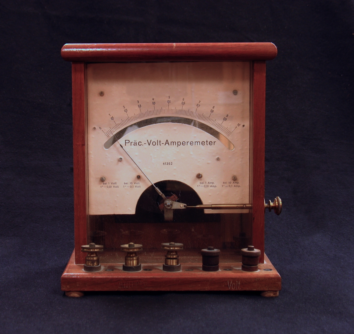 Ett av två stycken instrument för mätning av volt och ampere. "Låda" med botten-, sido-, topp- och bakstycke i trä. Genom en glasskiva syns en graderad mätare och nål.