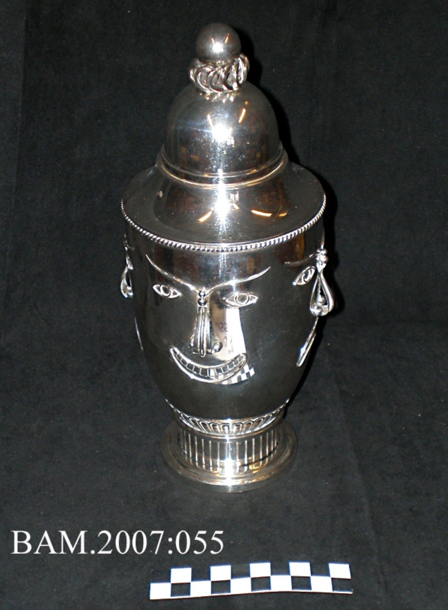 Sølvpokal med lokk. Stiliserte ansikter på "corpus". Utpreget urneform.