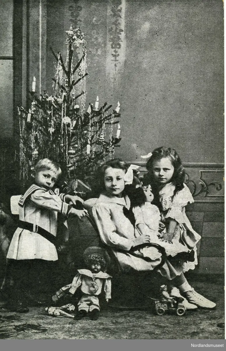 Postkortbilde av tre barn foran et pyntet juletre med lys inne i en stue. Gutt lengst til venstre har lyst hår og matrosdress, jente i midten sitter i en stol med en dukke på fanget og sløyfe i håret, jente lengst til høyre lener seg mot dukken. Foran dem står det enda en dukke og en lekebil med liten dukke i. 

Bakside: Oransje/rød postfrimerke, posthorn, 3 øre. Stemplet Dyrøhavn, 19.12.1908.