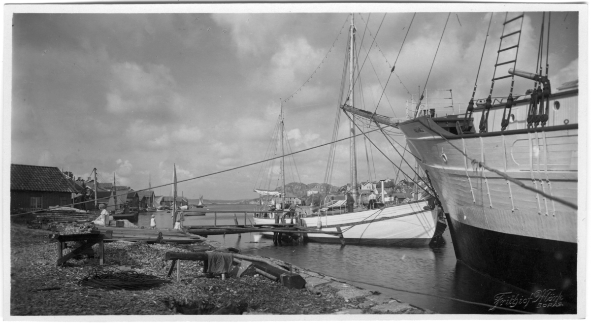 'Från Kilen, norrut. Båtar invid strand. 2 barn och 1 person på en hög träplankor på strand. På andra sidan hus på berghällar. ::  :: Serie fotonr 1888-1963.'