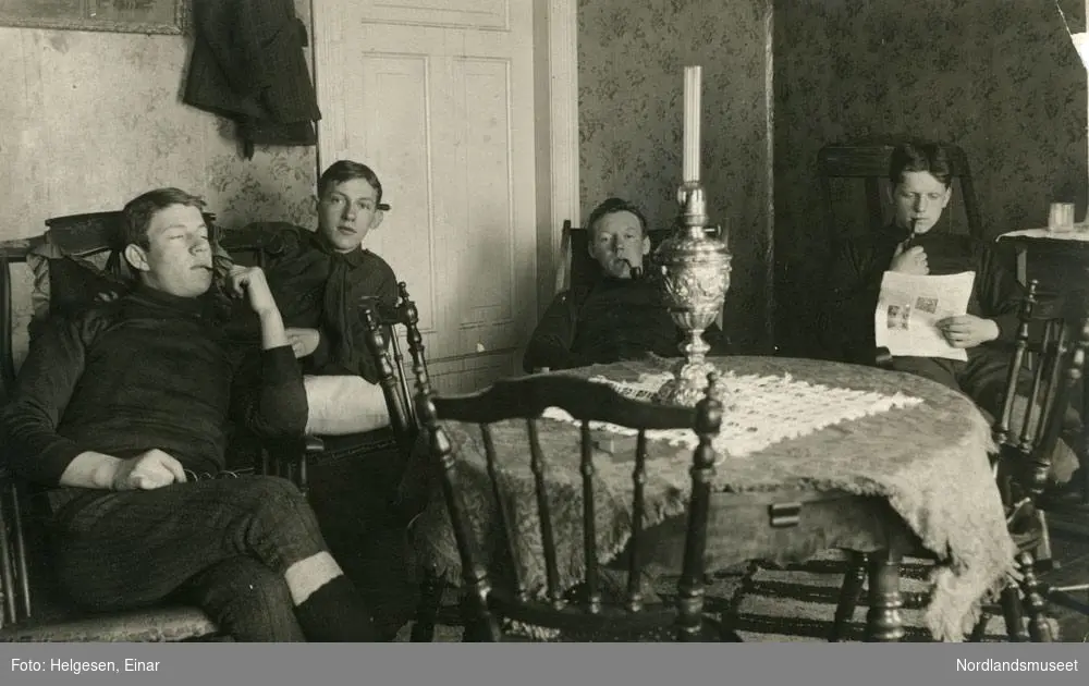 Påsketur til Vatnvatnet 1915. Henrik Andersen til høyre. Fire menn sitter i ei stue. Røyker pipe. Parafinlampe på bordet. En leser avisen.