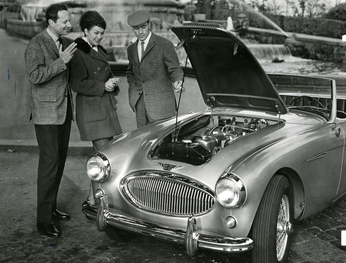 To menn og en kvinne røyker sigaretter og ser på en sportsbil av merket Austin.