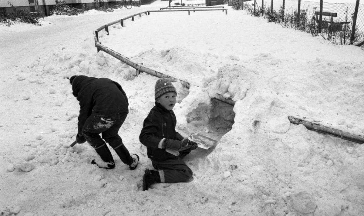 Snögrottor, 4 december 1965 

Man varnar för 'farliga snögrottor' i snöhögarna från snöplogarna. Två pojkar har skyfflat upp en hög med snö bakomstaketet runt gräsmattan. Nu håller den ene pojken på att gräva ur öppningen till grottan medan den andre skyfflar upp mer snö.