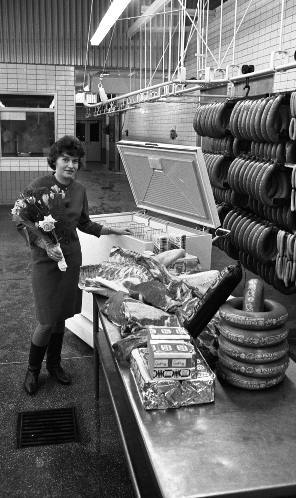 Orubricerat 18 februari 1966

Kvinna med en blombukett i sin vänstra hand står i en charkuterifabrik. Bredvid henne står en frysbox. I förgrunden står ett bord fyllt med leverpsatej, korvar och fläskkött. Till höger hänger korvar i olika storlekar längs med väggen.