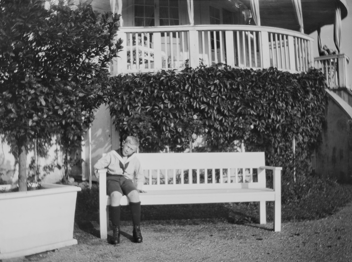 Iacob Ihlen Mathiesen sitter på en benk og myser mot sommersola. Han har skjorte med matroskrage. Bak ham en rund veranda dekket av klatreplanter.