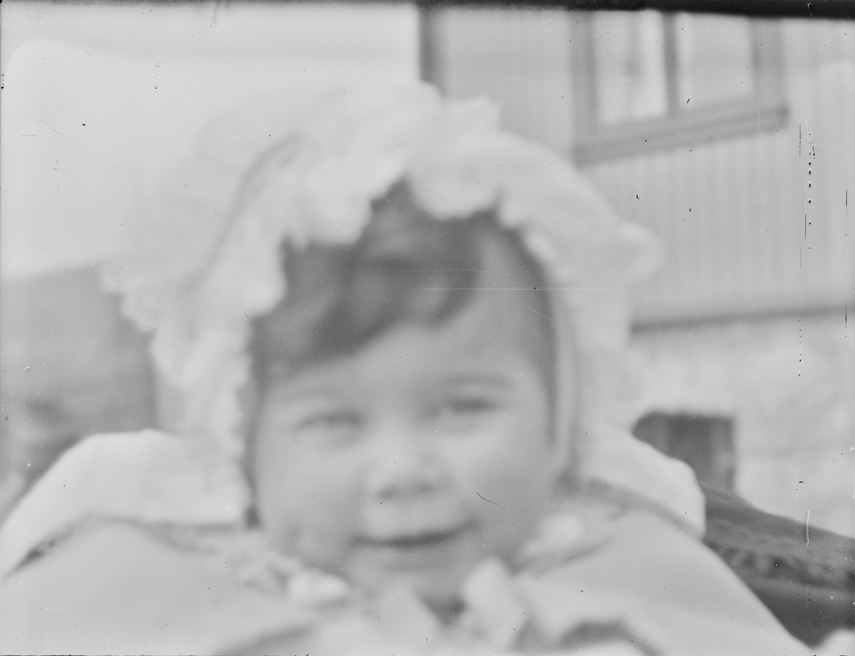 Nærbilde av et barn som sitter i en barnevogn utenfor et hus. Barnet smiler lurt under en pannelugg av mørke krøller.