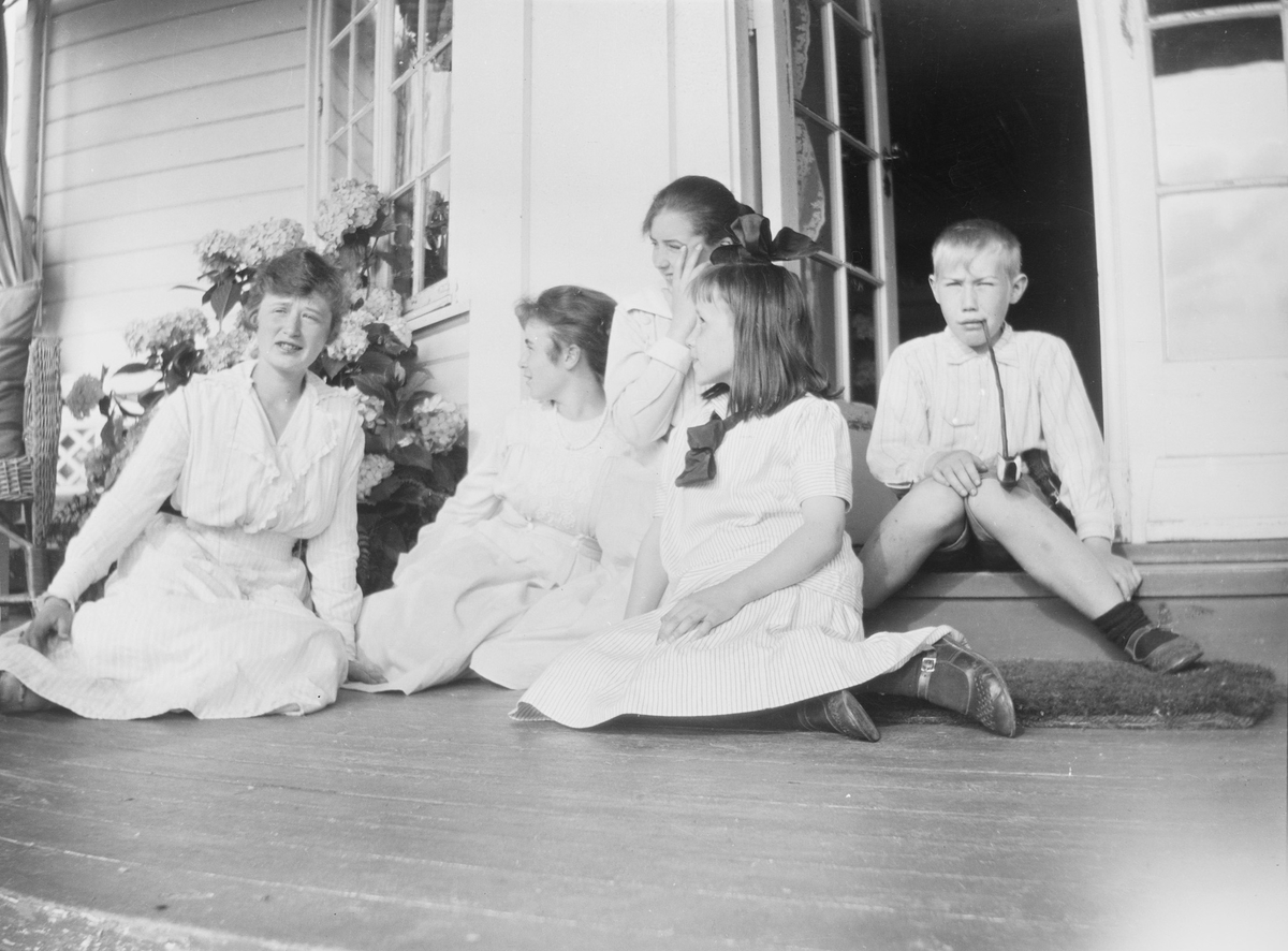 Fire kvinner, fra jente til voksen, sitter på verandagulvet sammen med Iacob som sitter på et trappetrinn.