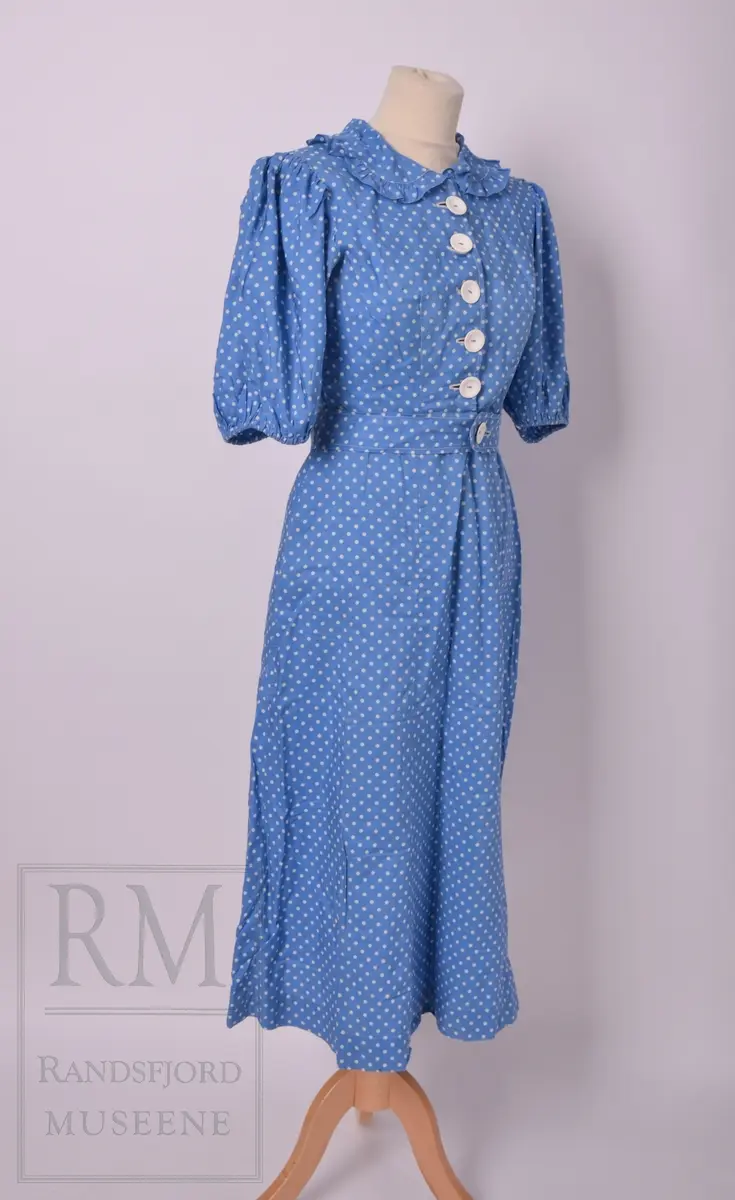Sommerkjole m/hatt. a) Bomullstoff, blått med hvite prikker. Sammensydd i livet. Overdelen har Lukning foran med 5 hvite knapper og knapphull, puffermer. Krage med heppekant. 6 Bredder i utskrådd skjørt. Lukning til venstre for midt foran. 
b) Belte med knapp og knapphull. 
c) Hatt, Heklet av hvitt bomullsgarn. Ståltråd ytterst i Bremmen (rusten) Figurkant på pullen. Pyntebånd av kjoletøyet.